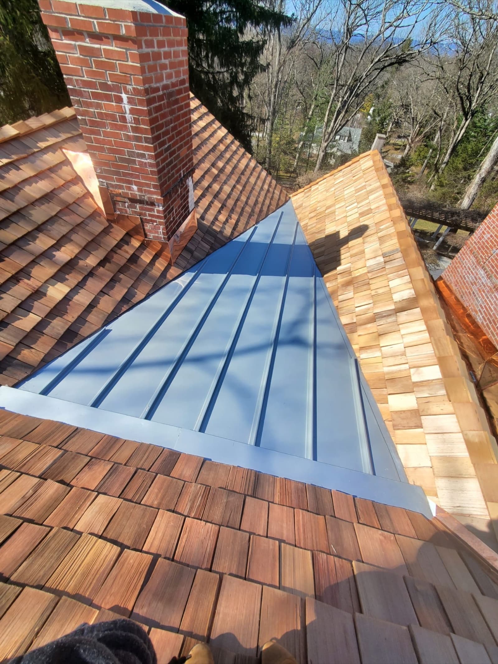 Standing-seam metal valley between cedar-shake slopes with a brick chimney — Zubar Roofing custom installation.